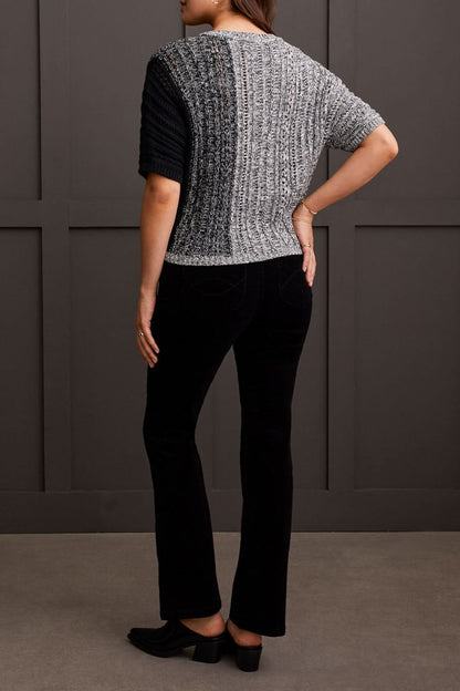 A person wearing the Tribal Dolman Elbow Sleeve Sweater, a cozy dual-toned knit featuring black and gray sections, short Dolman elbow sleeves, and a V-neckline, is paired with black pants and stands against a dark background.