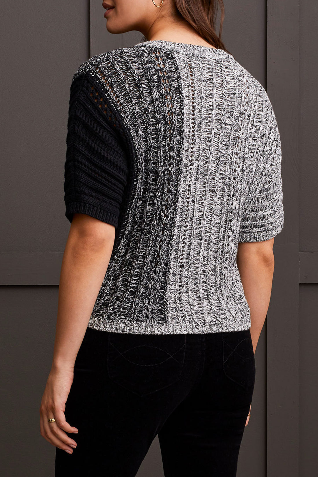 A person wearing the Tribal Dolman Elbow Sleeve Sweater, a cozy dual-toned knit featuring black and gray sections, short Dolman elbow sleeves, and a V-neckline, is paired with black pants and stands against a dark background.