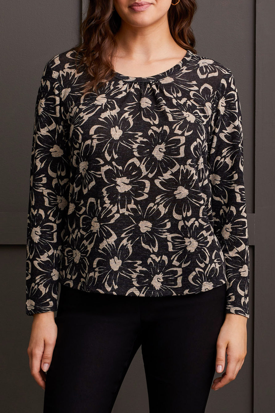A person stands wearing the U Neck Top with Shirring from Tribal, a flattering fit black long-sleeve top with a beige floral pattern, paired with black pants.