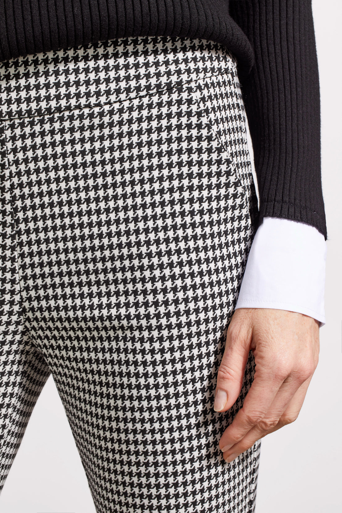 Close-up of a person showcasing the Tribal Houndstooth Pull on Pant, blending comfort and style with its classic houndstooth pattern. They're paired with a black sweater over a white cuffed shirt, revealing part of the torso and one hand.