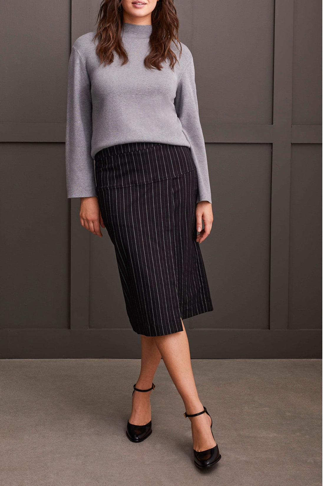 A person wearing a grey long-sleeve top and a black pinstripe pencil skirt with a slit, standing confidently with one hand on their hip, showcasing effortless style. This Striped Pull on Skirt by Tribal is a versatile addition to any wardrobe.