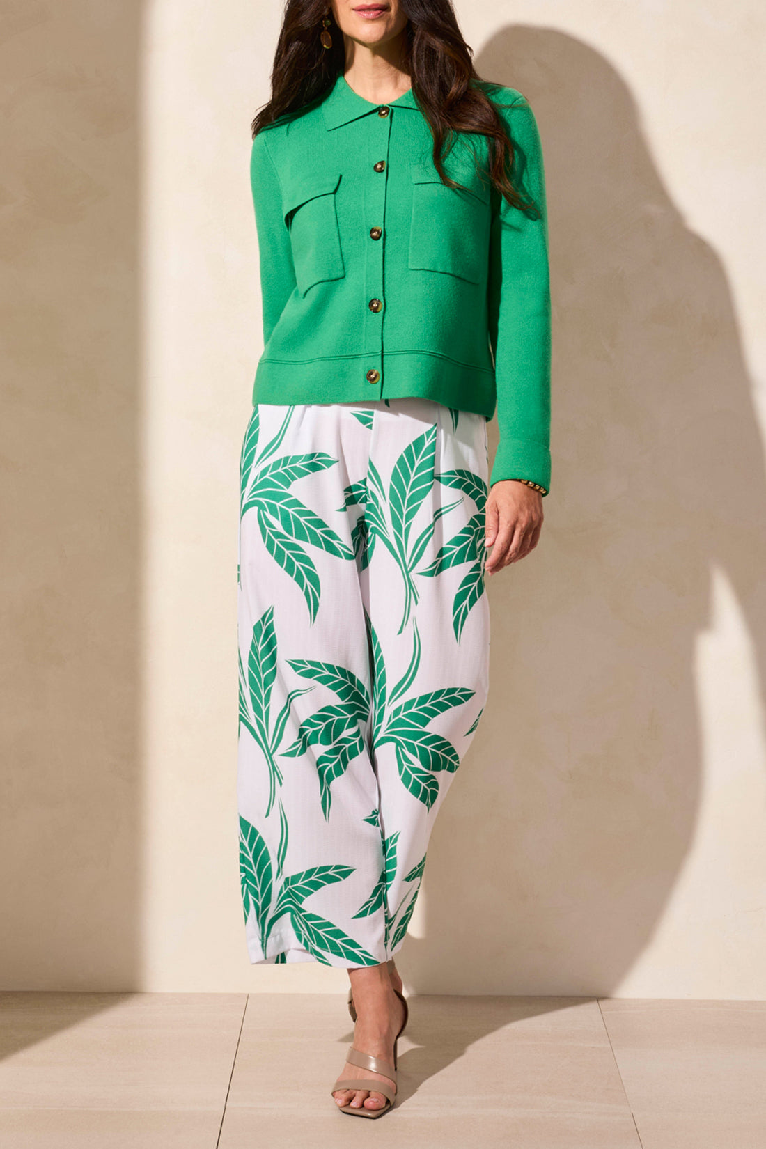 A person stands against a light wall wearing a Tribal DOUBLE KNIT COLLARED SWEATER CARDIGAN in green, accompanied by white and green leaf-patterned pants and beige sandals.