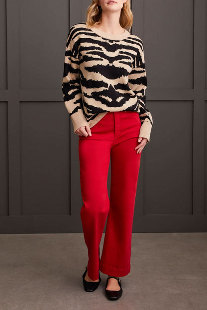 A person dressed in a chic and cozy Crew Neck Jacquard Sweater by Tribal, paired with red pants and black shoes, is standing against a paneled dark wall.