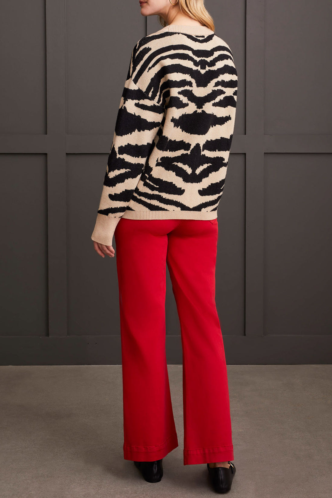 A person dressed in a chic and cozy Crew Neck Jacquard Sweater by Tribal, paired with red pants and black shoes, is standing against a paneled dark wall.