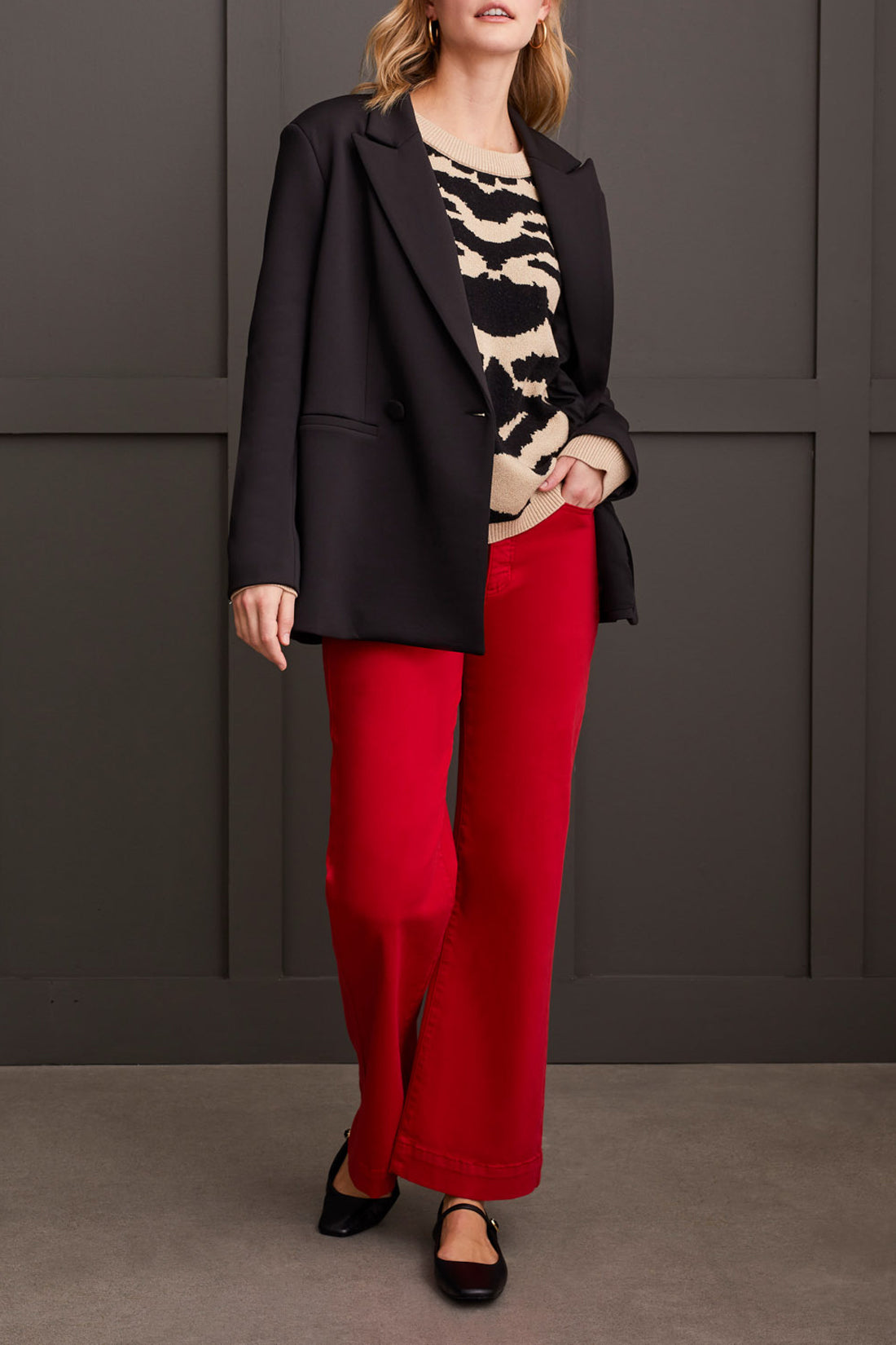 A person dressed in a chic and cozy Crew Neck Jacquard Sweater by Tribal, paired with red pants and black shoes, is standing against a paneled dark wall.