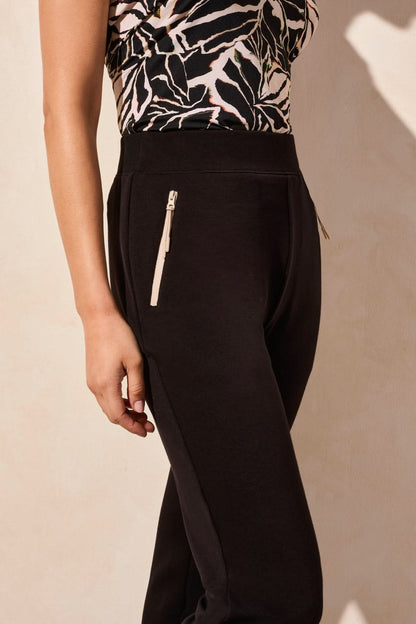 A person in Tribal's black PULL ON ANKLE PANT WITH POCKETS, paired with a patterned top and white sneakers, stands against a neutral background.