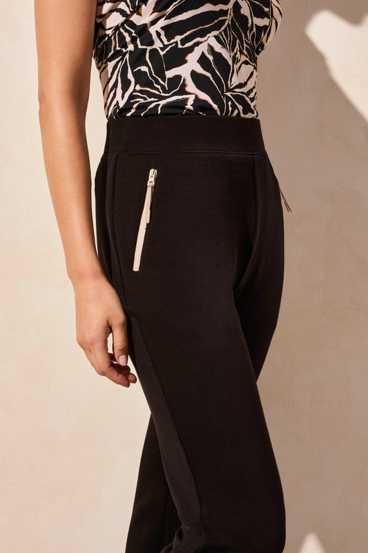 A person in Tribal's black PULL ON ANKLE PANT WITH POCKETS, paired with a patterned top and white sneakers, stands against a neutral background.