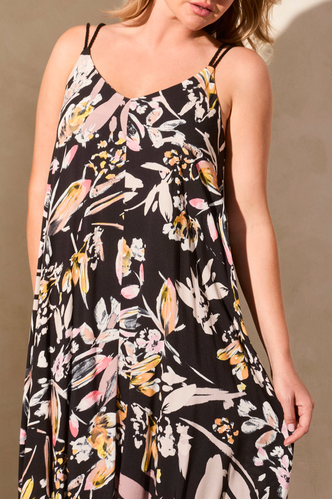 A person stands against a light wall wearing the Tribal brand LINED MAXI DRESS WITH ADJUSTABLE CORDS. The sleeveless black floral dress features a V-neckline and offers elegance and comfort for sunny days, with hands in pockets and paired with wedge sandals.