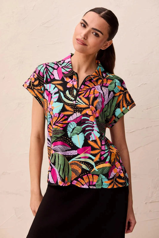 A woman wearing the Tribal HIGH PERFORMANCE DOLMAN TOP featuring a colorful tropical leaf pattern stands against a light textured wall.