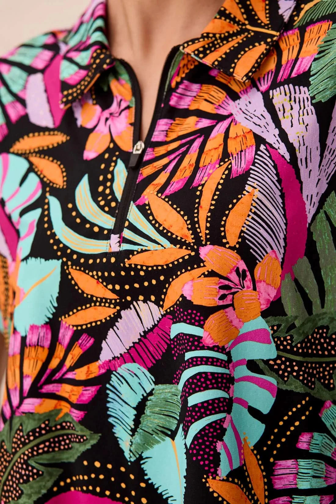 A woman wearing the Tribal HIGH PERFORMANCE DOLMAN TOP featuring a colorful tropical leaf pattern stands against a light textured wall.