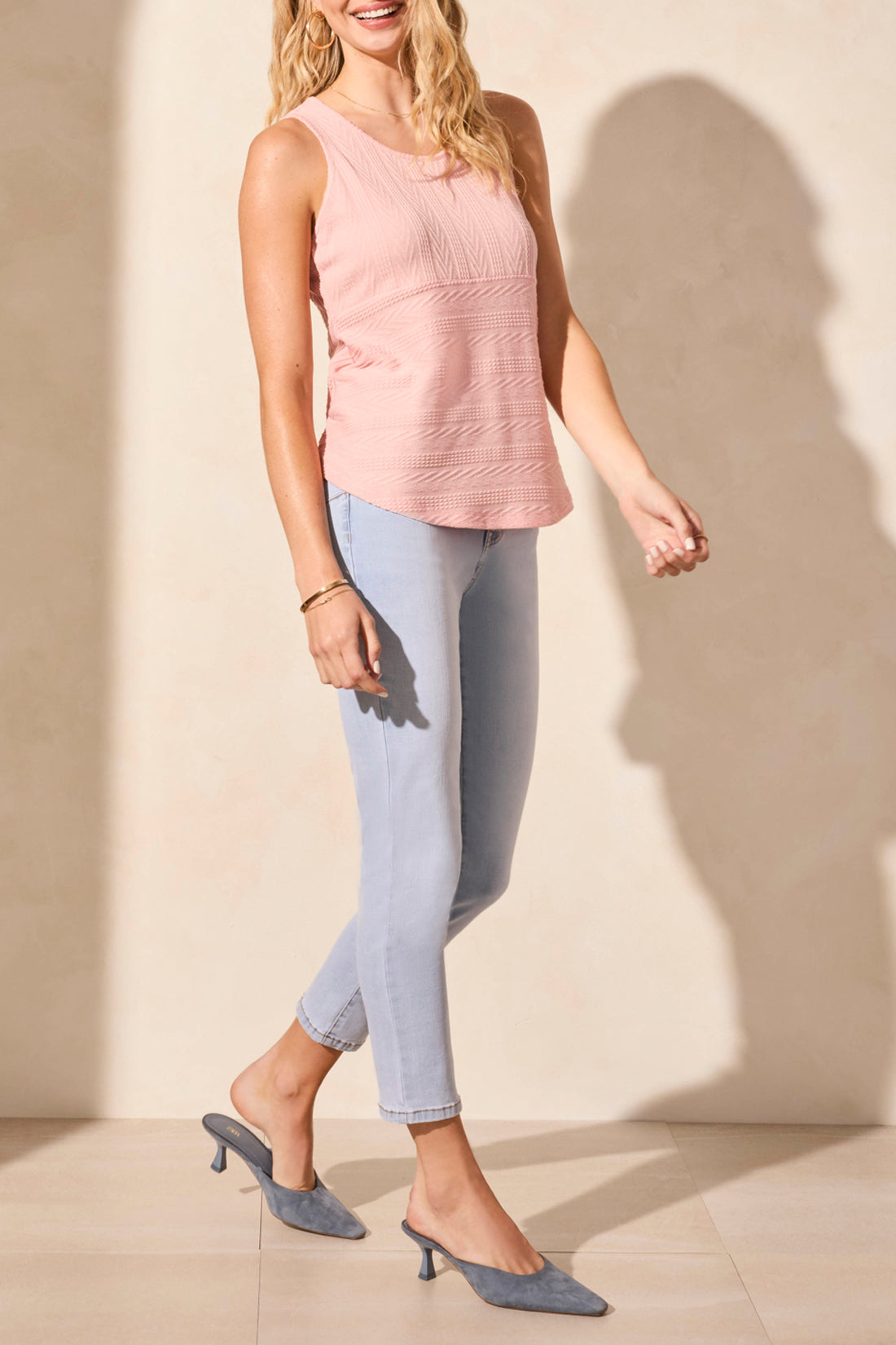 A person in a Tribal SLEEVELESS TOP WITH SHIRTTAIL, paired with light blue jeans, stands next to a wooden stool. The top is white and bra-friendly, perfect for the well-lit room setting.