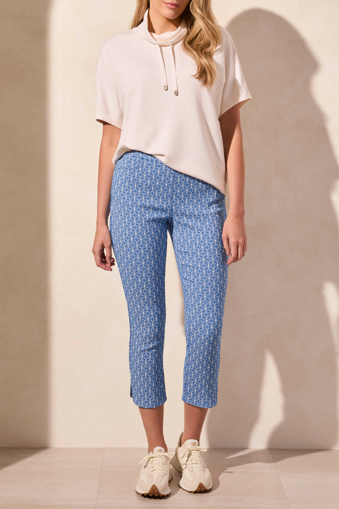 Wearing Tribal's SHORT SLEEVE FUNNEL NECK TOP in light beige, a person pairs it with blue patterned pants against a plain background. The versatile top features a flattering fit and drawstrings.