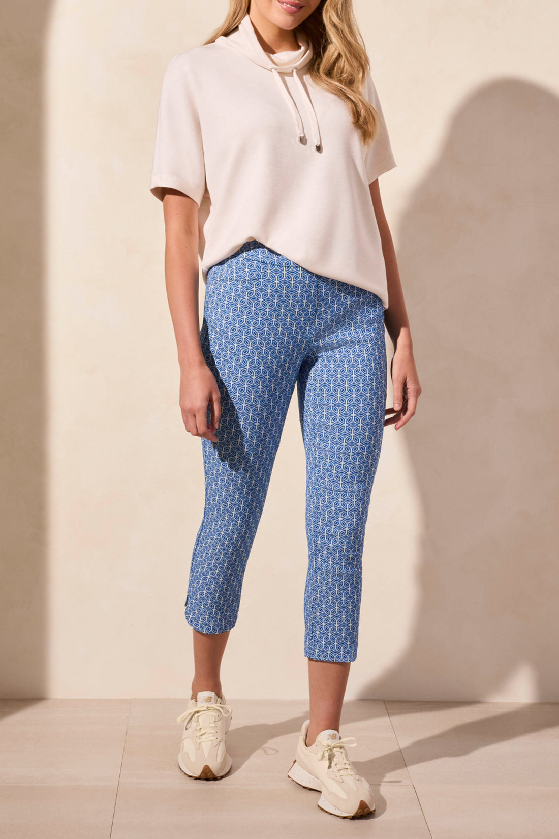 Wearing Tribal's SHORT SLEEVE FUNNEL NECK TOP in light beige, a person pairs it with blue patterned pants against a plain background. The versatile top features a flattering fit and drawstrings.