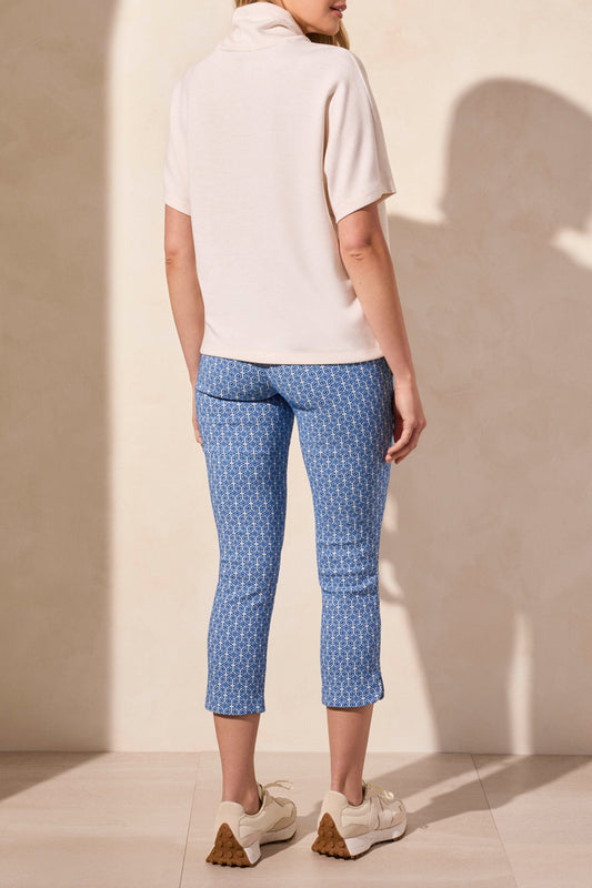 Wearing Tribal's SHORT SLEEVE FUNNEL NECK TOP in light beige, a person pairs it with blue patterned pants against a plain background. The versatile top features a flattering fit and drawstrings.
