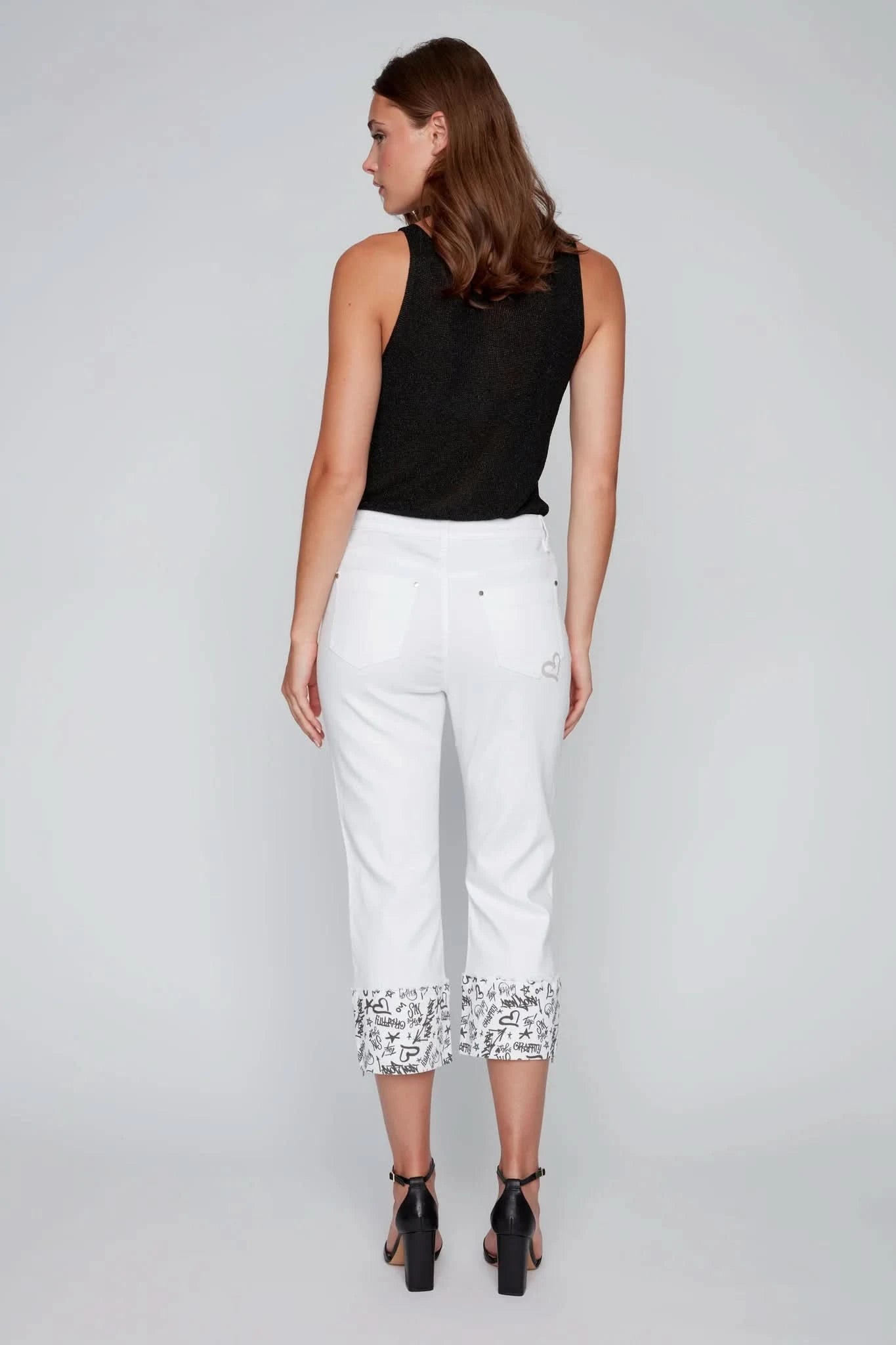 A woman poses in a studio wearing a black sleeveless top, CoCo Y Club’s White Capris with Graffiti Cuffs, and black heeled sandals—an ideal summer fashion look.