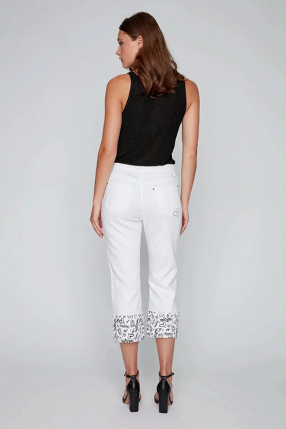 A woman poses in a studio wearing a black sleeveless top, CoCo Y Club’s White Capris with Graffiti Cuffs, and black heeled sandals—an ideal summer fashion look.