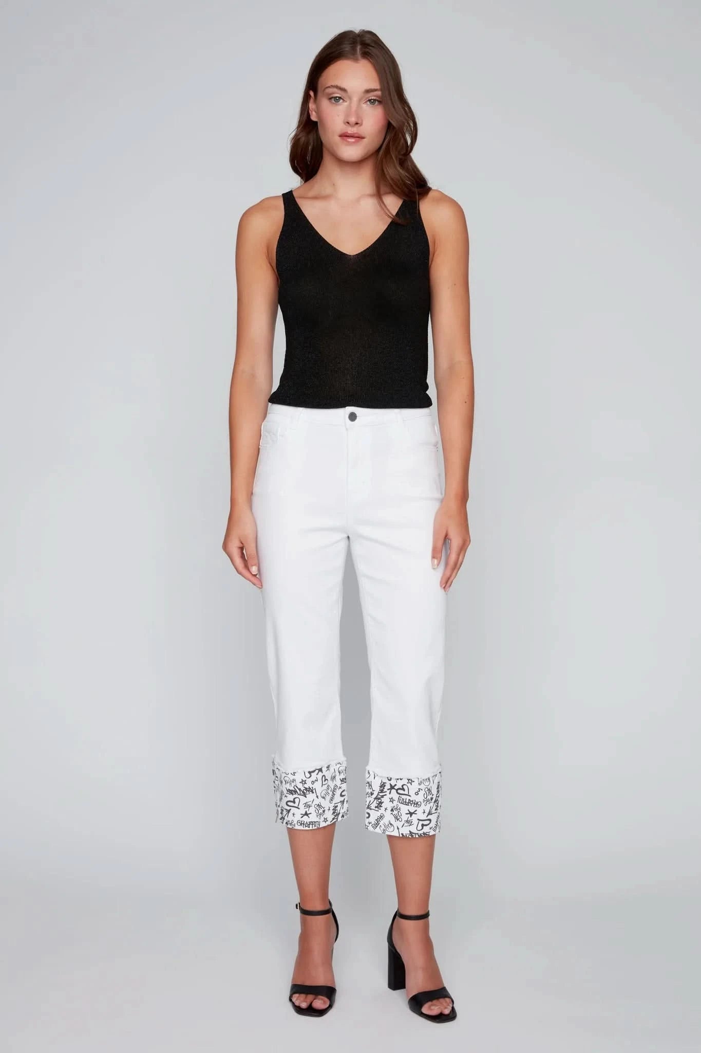 A woman poses in a studio wearing a black sleeveless top, CoCo Y Club’s White Capris with Graffiti Cuffs, and black heeled sandals—an ideal summer fashion look.