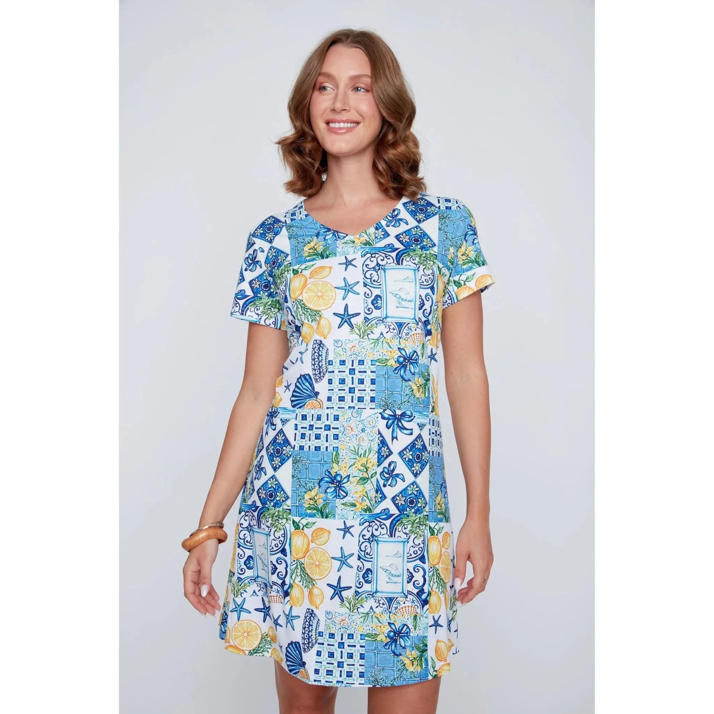 A woman stands smiling in the CoCo Y Club ‘Amalfi’ Lace Detail Dress, a short sleeve style featuring blue and yellow Greek-inspired tile and lemon motifs on a white background.