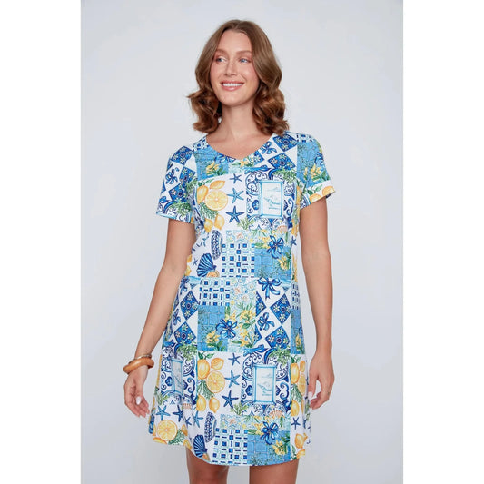 A woman stands smiling in the CoCo Y Club ‘Amalfi’ Lace Detail Dress, a short sleeve style featuring blue and yellow Greek-inspired tile and lemon motifs on a white background.
