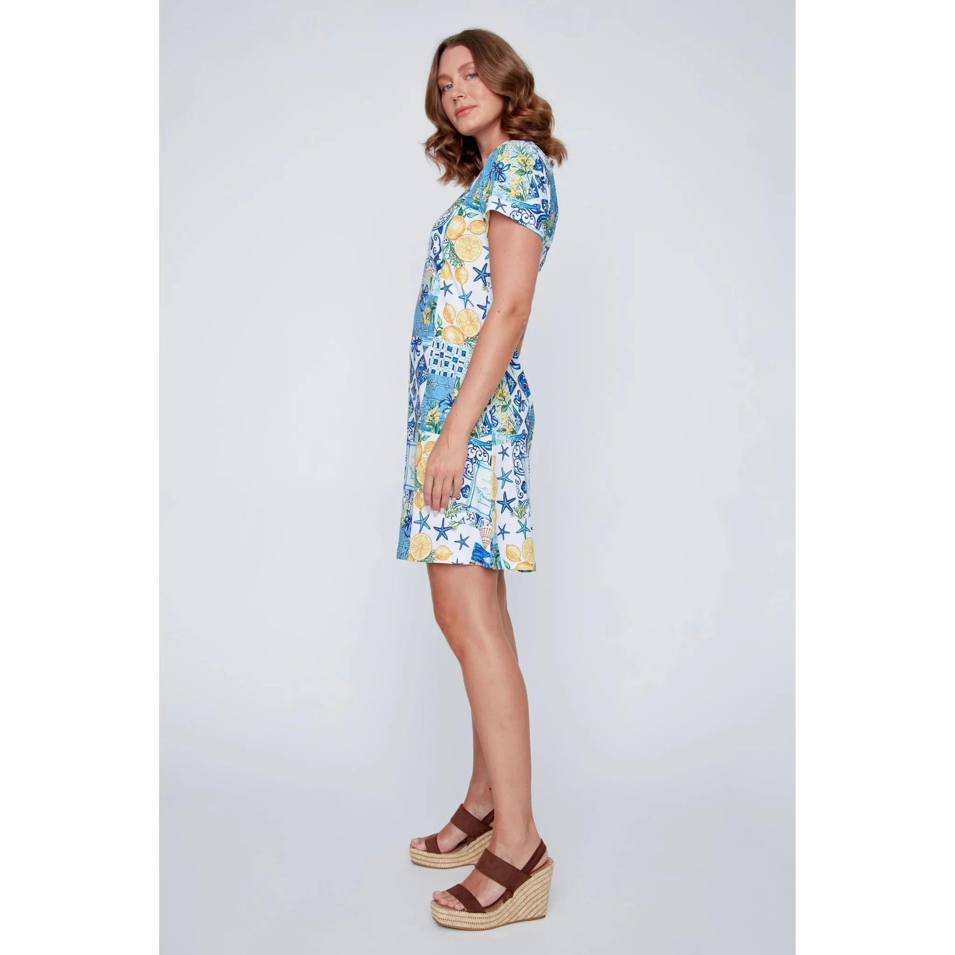 A woman stands smiling in the CoCo Y Club ‘Amalfi’ Lace Detail Dress, a short sleeve style featuring blue and yellow Greek-inspired tile and lemon motifs on a white background.