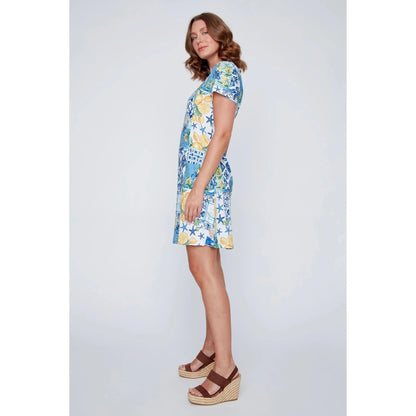 A woman stands smiling in the CoCo Y Club ‘Amalfi’ Lace Detail Dress, a short sleeve style featuring blue and yellow Greek-inspired tile and lemon motifs on a white background.