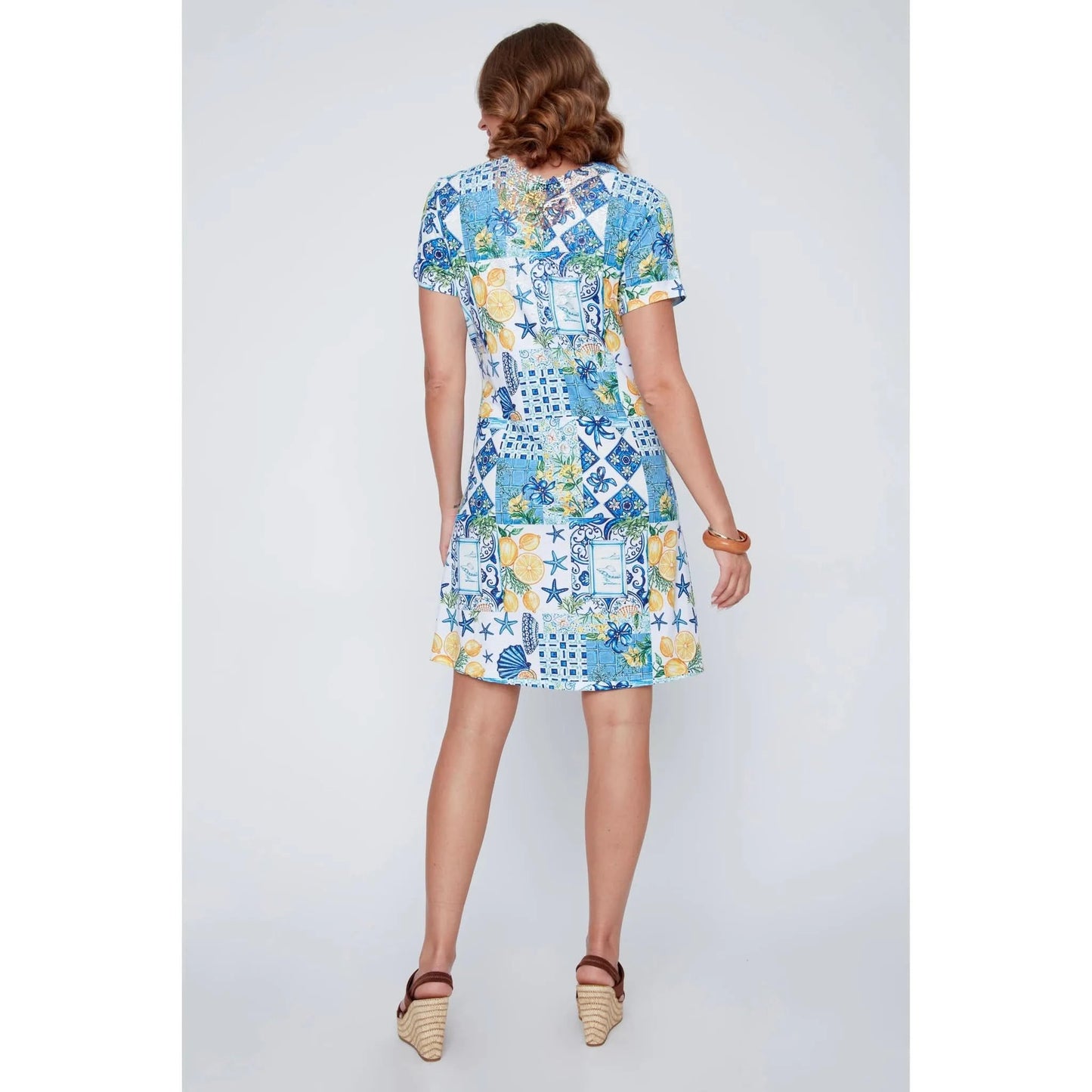 A woman stands smiling in the CoCo Y Club ‘Amalfi’ Lace Detail Dress, a short sleeve style featuring blue and yellow Greek-inspired tile and lemon motifs on a white background.