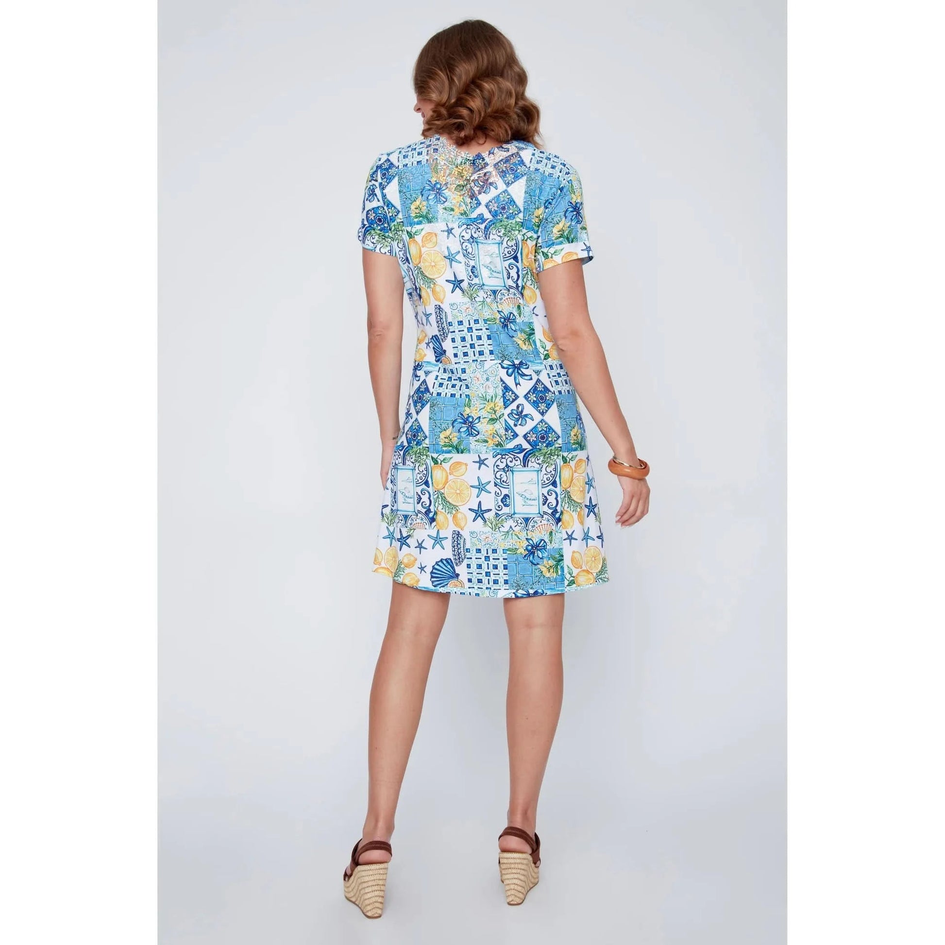 A woman stands smiling in the CoCo Y Club ‘Amalfi’ Lace Detail Dress, a short sleeve style featuring blue and yellow Greek-inspired tile and lemon motifs on a white background.