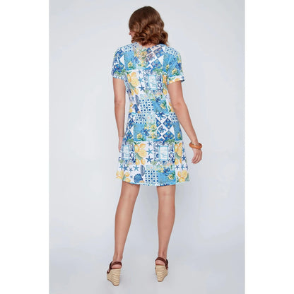 A woman stands smiling in the CoCo Y Club ‘Amalfi’ Lace Detail Dress, a short sleeve style featuring blue and yellow Greek-inspired tile and lemon motifs on a white background.