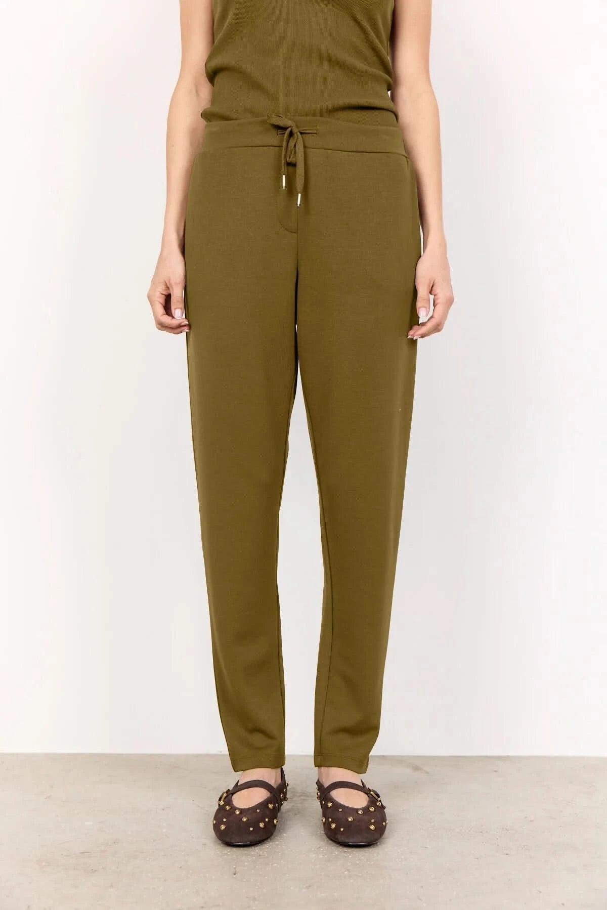 A woman stands against a white wall wearing Soya Concepts Dark Olive Sweatpants with an adjustable waist, hands in pockets, paired with a sleeveless top and brown studded flats.
