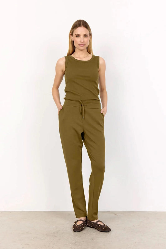 A woman stands against a white wall wearing Soya Concepts Dark Olive Sweatpants with an adjustable waist, hands in pockets, paired with a sleeveless top and brown studded flats.