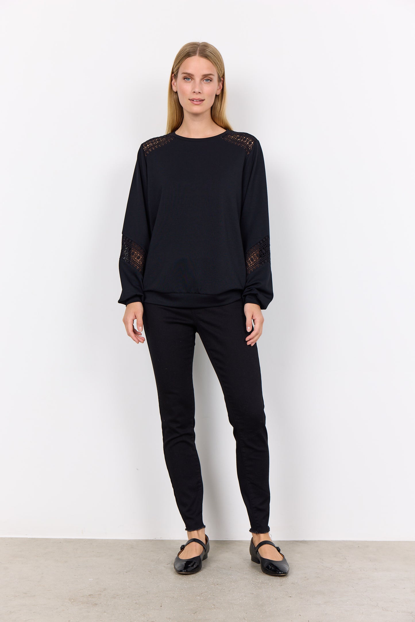 A woman wearing the Soya Concepts BANU 193 Sweater, featuring intricate lace and embroidery details on the shoulders and sleeves, stands against a plain white background. She has long, light brown hair and is looking at the camera, exuding a stylish yet comfortable fit.