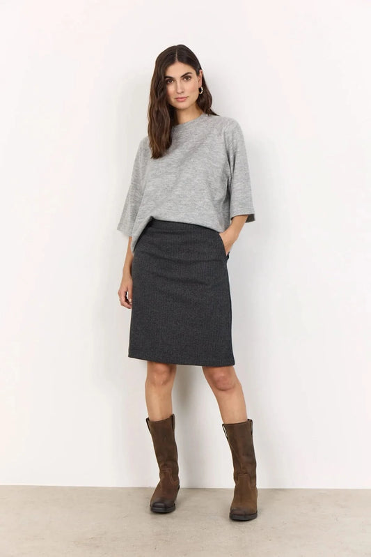 A woman stands against a white wall, wearing a Soya Concepts Short Sleeve Light Grey Knitted Sweater, a dark knee-length skirt, and brown knee-high boots. One hand is in her skirt pocket.