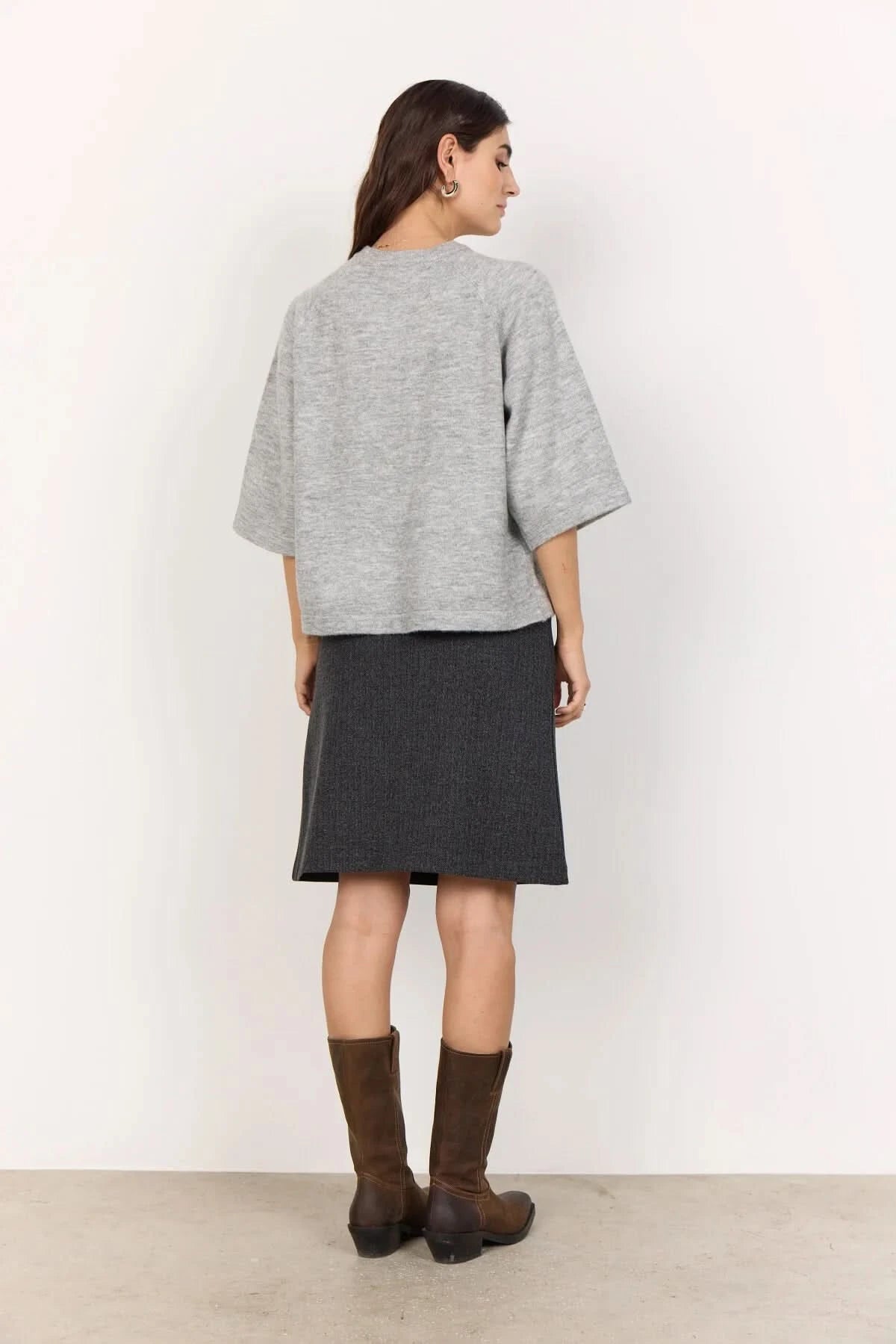 A woman stands against a white wall, wearing a Soya Concepts Short Sleeve Light Grey Knitted Sweater, a dark knee-length skirt, and brown knee-high boots. One hand is in her skirt pocket.