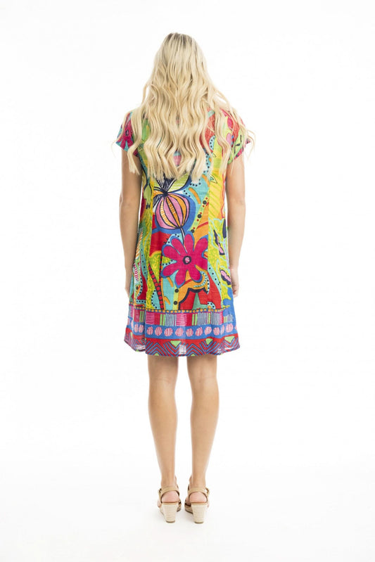 A woman stands against a white background wearing the Orientique San Miguel Bib Dress with Pockets, made from vibrant cotton slub with a pleated bust detail, paired with beige sandals.