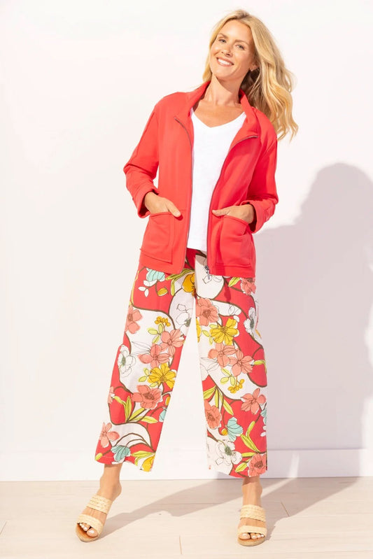 A woman stands against a white background in Escape’s Seaside knit jacket—a vibrant red layer over a white top, paired with floral wide-leg pants and sandals—ideal for transitioning through changing coastal weather.
