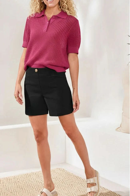 A woman stands indoors wearing a textured short-sleeve magenta polo shirt, Tribal FLATTEN IT SHORTS with a button, and white sandals.