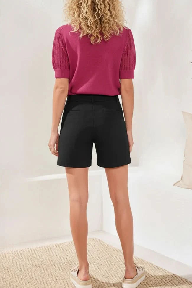 A woman stands indoors wearing a textured short-sleeve magenta polo shirt, Tribal FLATTEN IT SHORTS with a button, and white sandals.