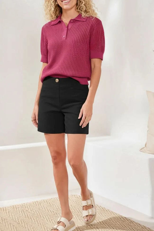 A woman stands indoors wearing a textured short-sleeve magenta polo shirt, Tribal FLATTEN IT SHORTS with a button, and white sandals.