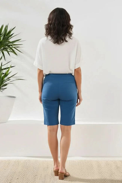 A person models the Tribal FLATTEN IT BERMUDA SHORTS in white with a pull-on waistband, paired with a white top, standing indoors near a plant, showing a side and partial back view.
