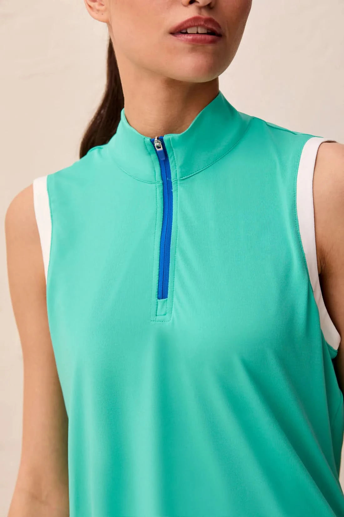 A woman models the Tribal SLEEVELESS ZIP SPORT TOP in teal, featuring a blue zippered collar and white armhole trim in lightweight scuba knit, standing indoors against a light-colored wall.