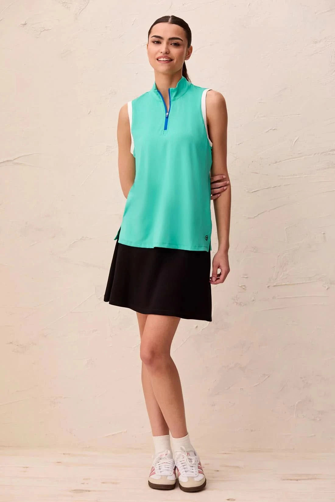 A woman models the Tribal SLEEVELESS ZIP SPORT TOP in teal, featuring a blue zippered collar and white armhole trim in lightweight scuba knit, standing indoors against a light-colored wall.
