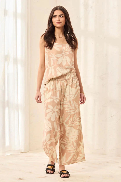 A woman models Tribal’s PULL ON CAPRI W/ WAISTBAND RUFFLE—sleeveless beige top and matching wide-leg capris with a subtle crosshatch pattern—standing indoors on a light floor with a soft curtain backdrop.