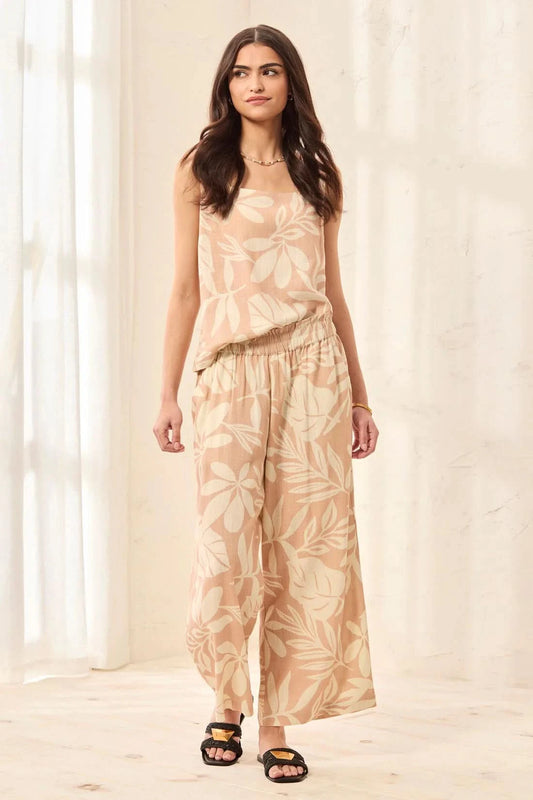 A woman models Tribal’s PULL ON CAPRI W/ WAISTBAND RUFFLE—sleeveless beige top and matching wide-leg capris with a subtle crosshatch pattern—standing indoors on a light floor with a soft curtain backdrop.
