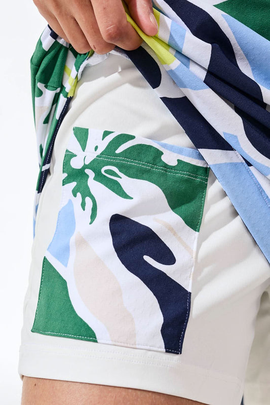 A woman in a white jacket and Tribal HIGH PERFORMANCE PULL-ON SKORT, with a visor and golf glove, stands on a golf course holding a club amid palm trees and buildings.