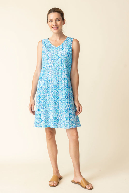 A woman smiles with her hair tied back, wearing Habitat’s Easy Pocket Dress—a sleeveless V-neck blue patterned dress with pockets—and tan flat sandals against a plain background.