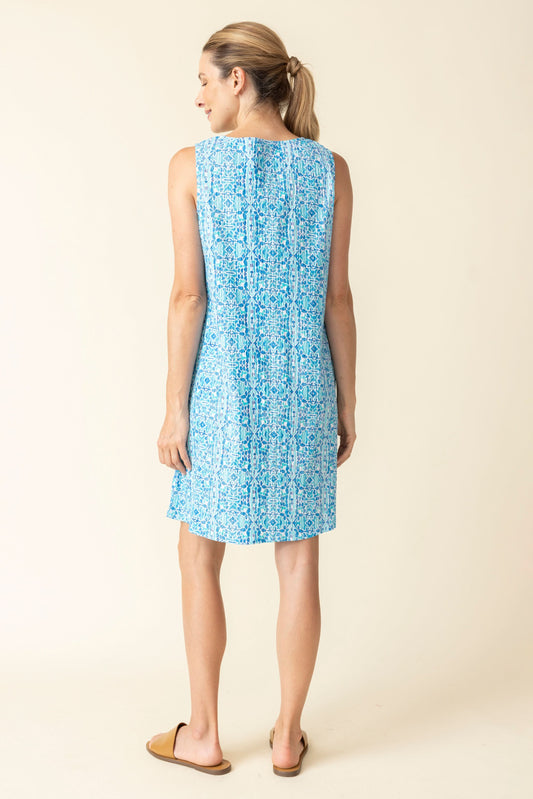 A woman smiles with her hair tied back, wearing Habitat’s Easy Pocket Dress—a sleeveless V-neck blue patterned dress with pockets—and tan flat sandals against a plain background.