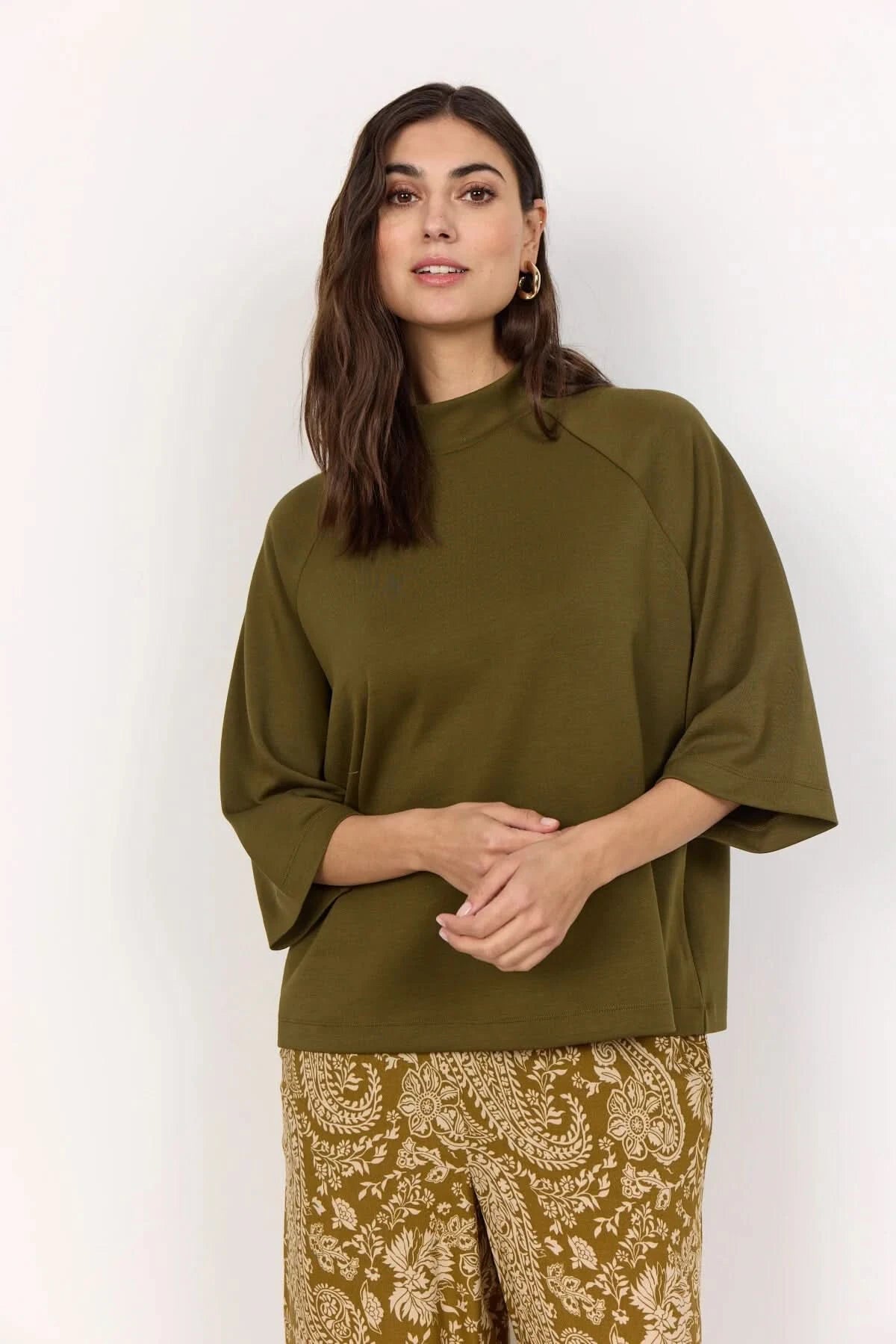 A woman stands against a plain light background wearing the Soya Concepts Short Sleeve Knit in dark olive, paired with brown paisley-patterned pants.