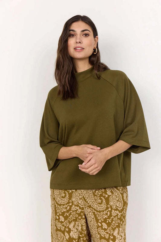 A woman stands against a plain light background wearing the Soya Concepts Short Sleeve Knit in dark olive, paired with brown paisley-patterned pants.