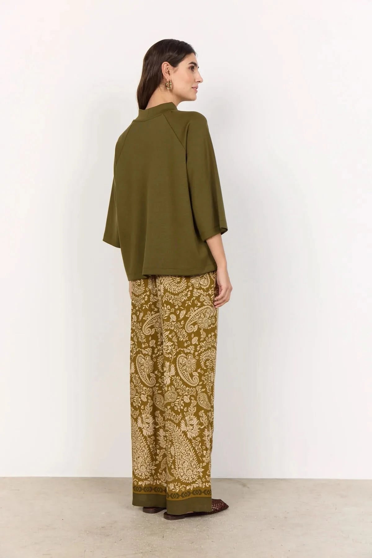 A woman stands against a plain light background wearing the Soya Concepts Short Sleeve Knit in dark olive, paired with brown paisley-patterned pants.