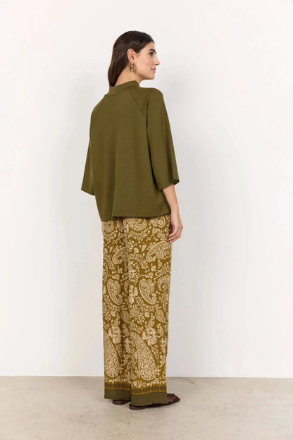 A woman stands against a plain light background wearing the Soya Concepts Short Sleeve Knit in dark olive, paired with brown paisley-patterned pants.
